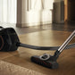 Miele Boost CX1 Cat & Dog - Obsidian Black — Sleek canister vacuum on hardwood floor with hose and brush head, cleaning near a rug in a sunlit modern room.