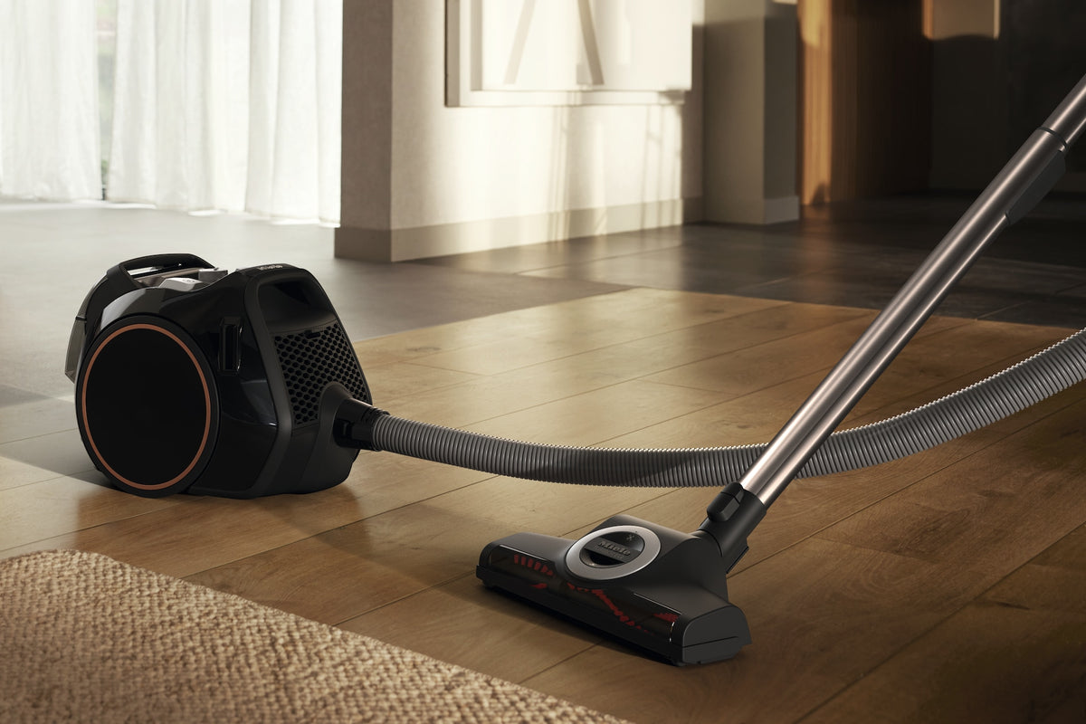 Miele Boost CX1 Cat & Dog - Obsidian Black — Sleek canister vacuum on hardwood floor with hose and brush head, cleaning near a rug in a sunlit modern room.