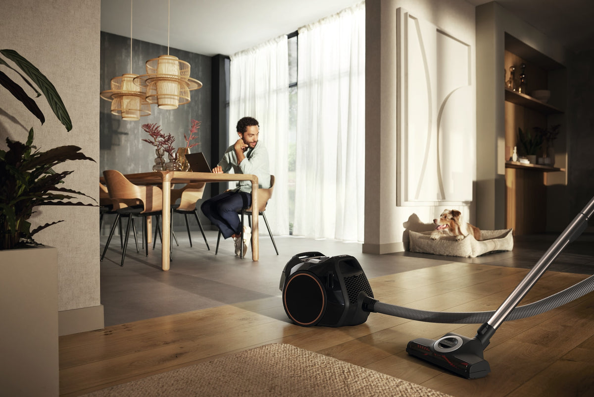 Miele Boost CX1 Cat & Dog - Obsidian Black — Modern living space with canister vacuum cleaning hardwood; person works at table, dog relaxing on bed.